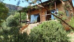 A rustic brick house with a wooden porch, surrounded by lush greenery and a mountainous backdrop. - Thirsty Falls Guest Farm (Maanhaarand)