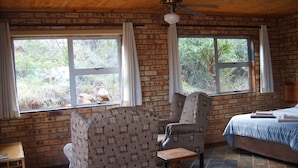 A room with a brick wall, two windows with curtains, a bed with a blue bedspread, a wooden table, and a patterned armchair. - Thirsty Falls Guest Farm (Maanhaarand)