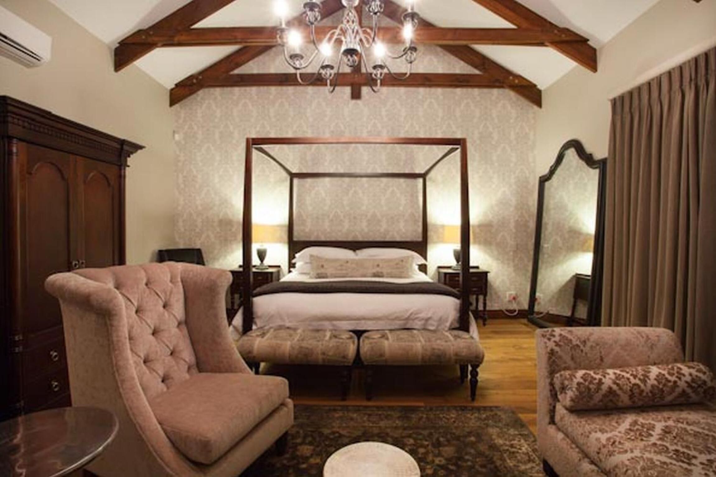 Un dormitorio con cama con dosel, una zona de estar con una silla y reposapiés, una mesa redonda y un espejo grande.