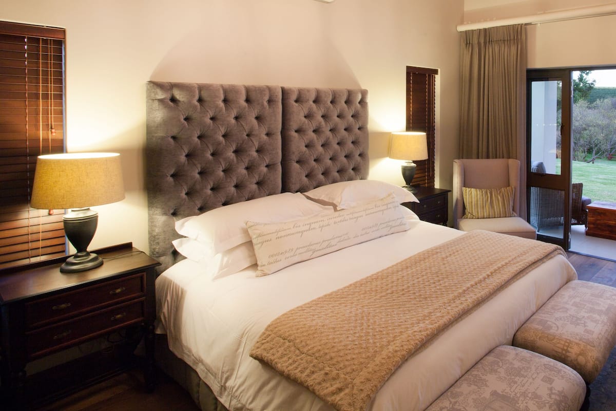A bedroom with a large bed, a nightstand, a lamp, a chair, and a view of a garden through a glass door.