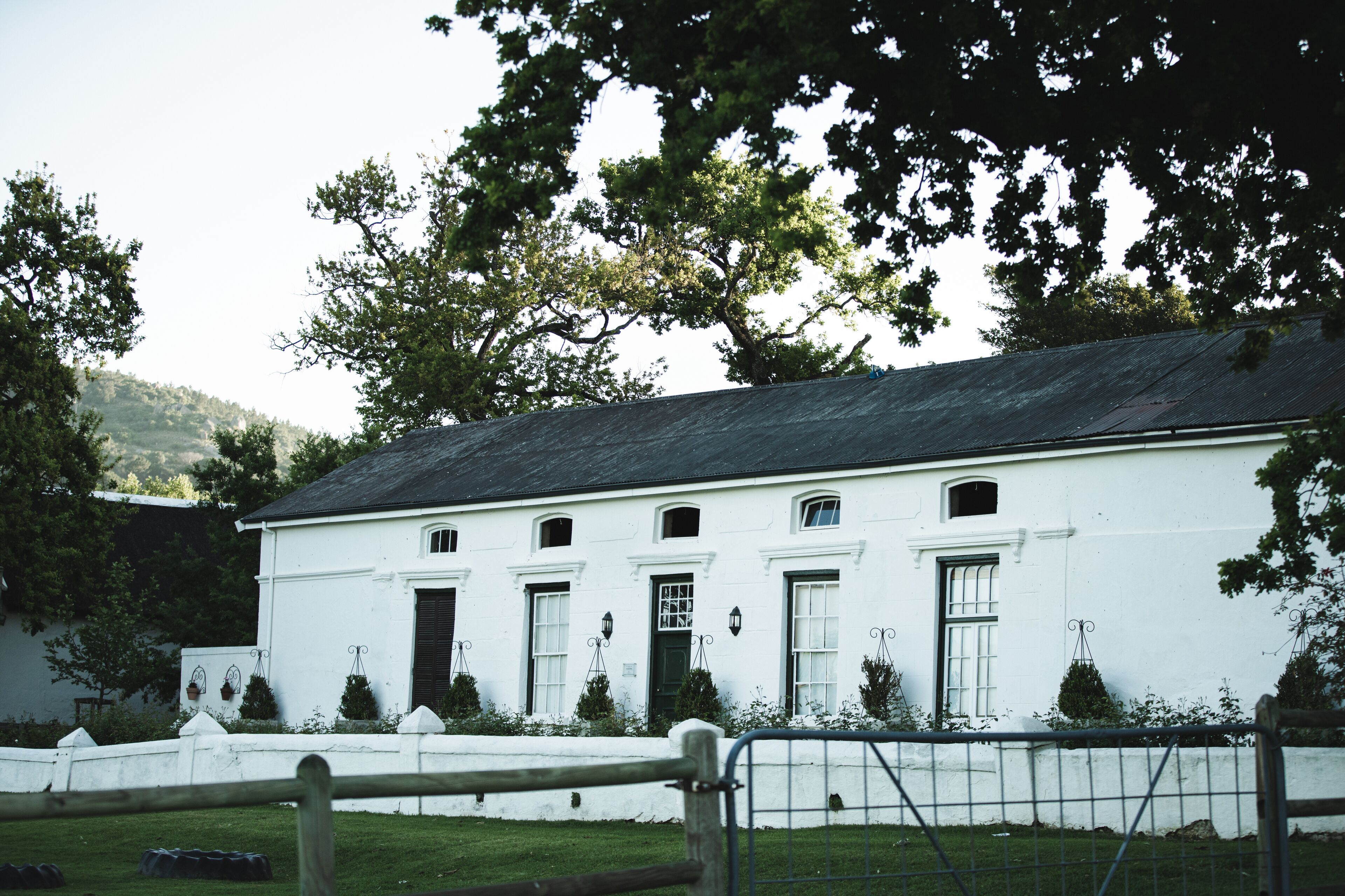 A white building with multiple windows and doors, surrounded by a white fence and greenery.