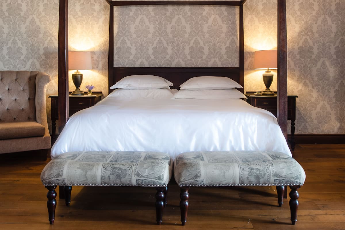 A four-poster bed with white linens, two bedside tables with lamps, and a tufted armchair.