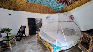 A room with a bed under a mosquito net, a chair, a small table with a plant, and a patterned bench. - Red Rocks Rwanda Guesthouse & Campsite (Nkotsi)