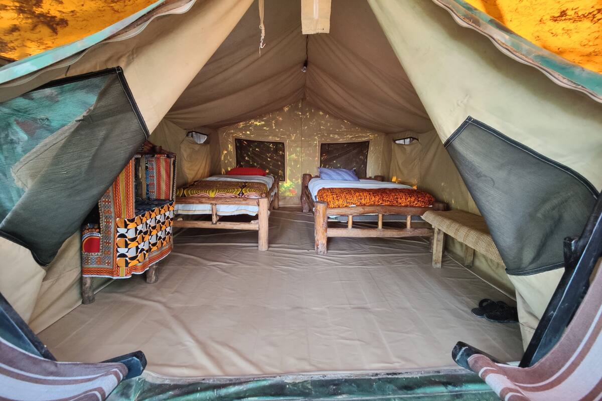 A tent with two beds, each with a wooden frame and a mattress. There is a small table with a colorful cloth between the beds.