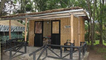A rustic wooden structure with a bamboo exterior, a sign reading "RUGAN," and a stone-paved entrance area.
