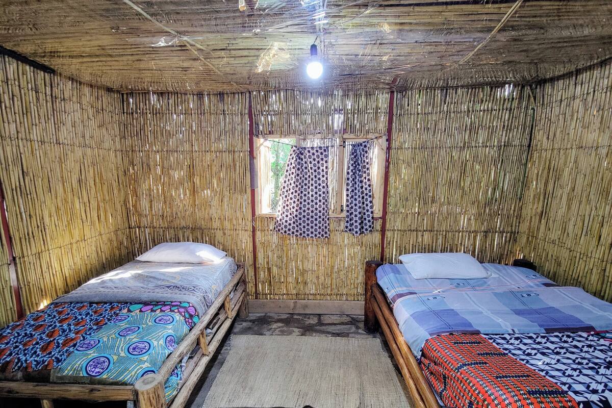 A room with two wooden beds, a bamboo wall, a small window with patterned curtains, and a stone floor.