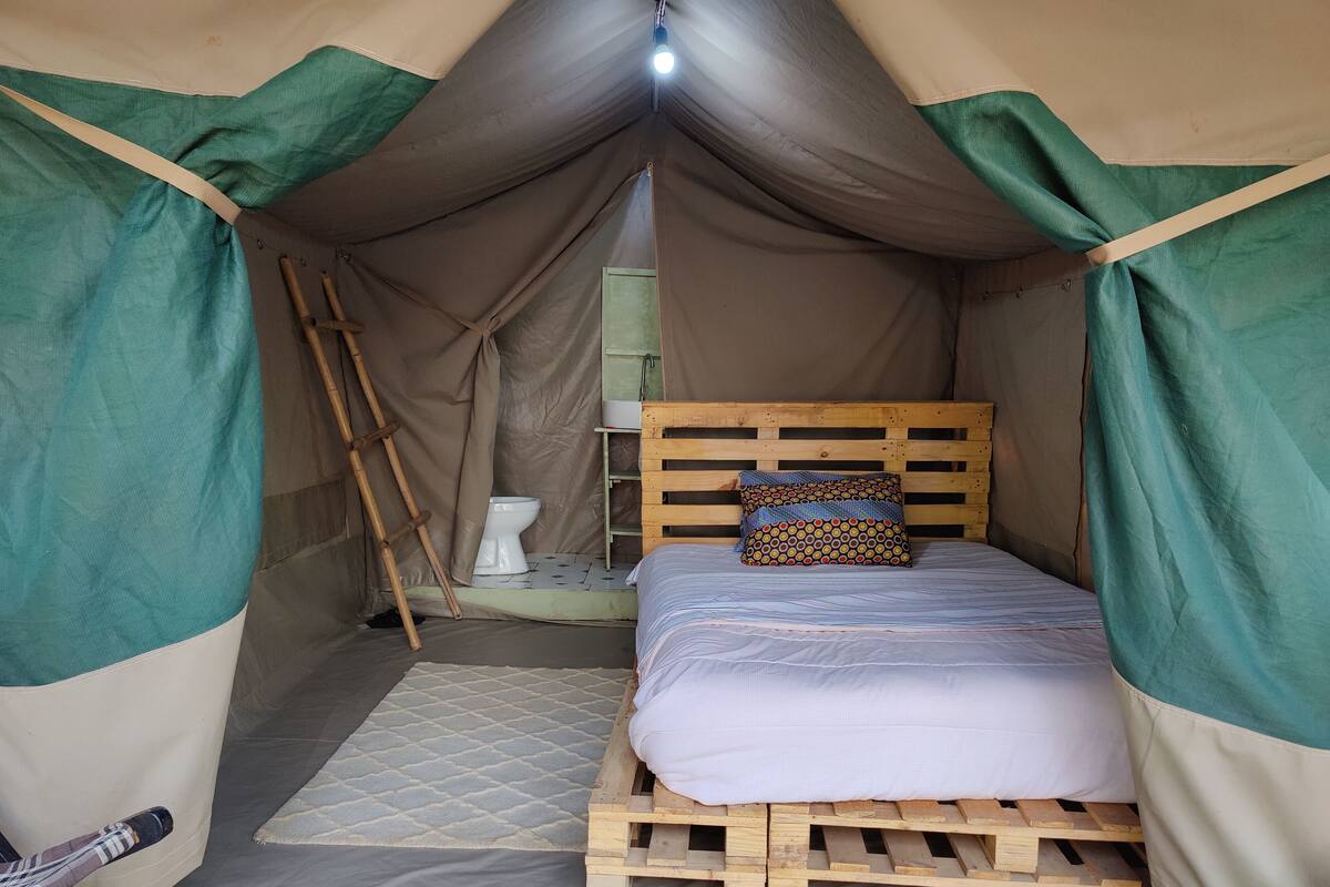 A tent with a bed, a wooden headboard, a pillow, a toilet, and a ladder.