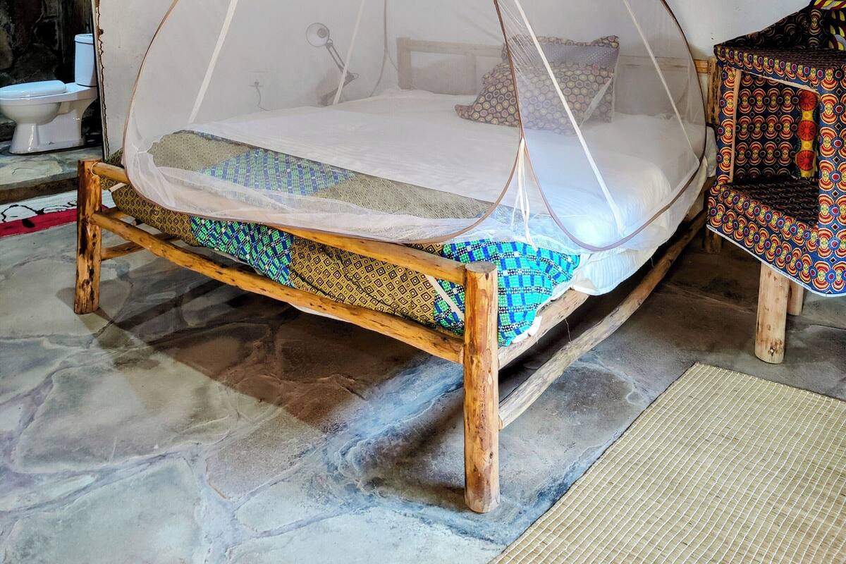 A room with a bed under a thatched roof, a painting on the wall, and a patterned rug on the floor.