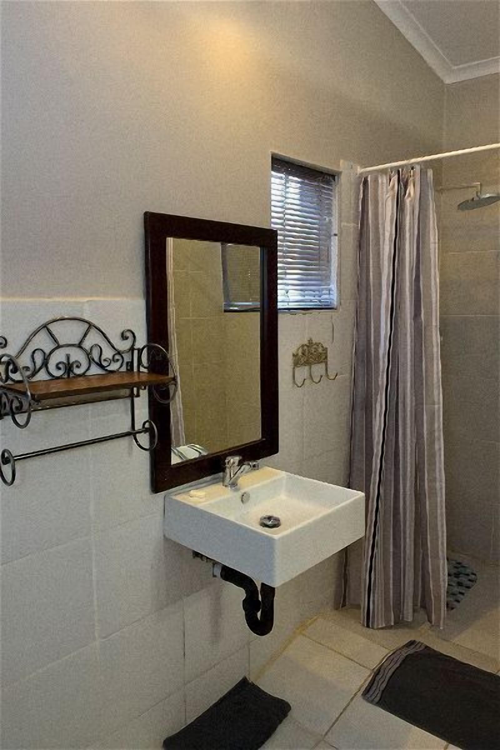 Un baño con lavamanos blanco, espejo, cortina de ducha y ventana con persianas.