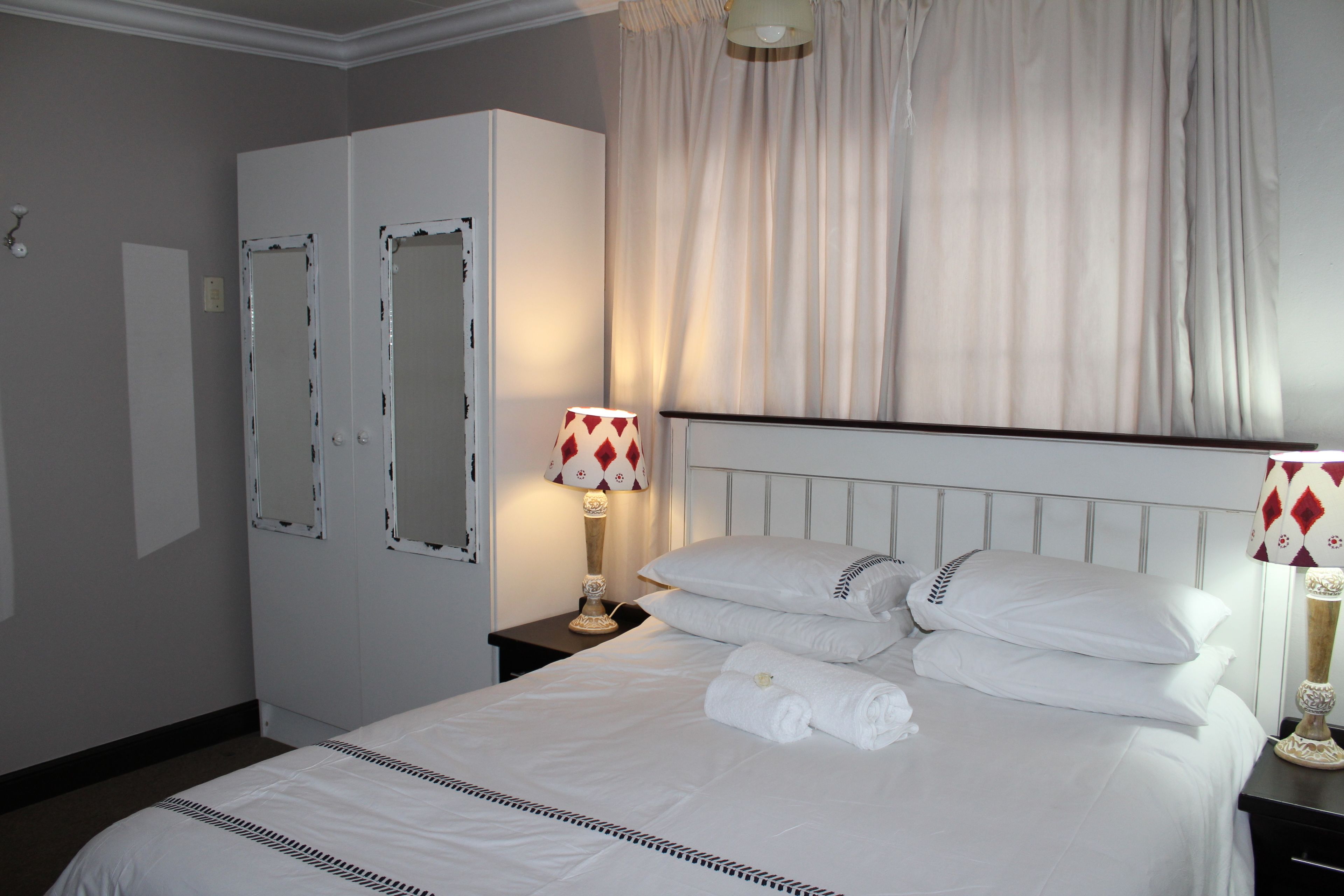 En pænt redt seng med hvide sengetøj og puder, et natbord med en lampe, og en garderobe med et spejl.