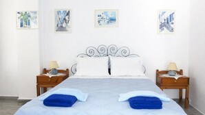 A double bed with a white and blue bedspread, two pillows, two blue towels, and a metal headboard. There are two bedside tables with lamps and framed pictures on the wall above. - Bungalows Marina (Paros)