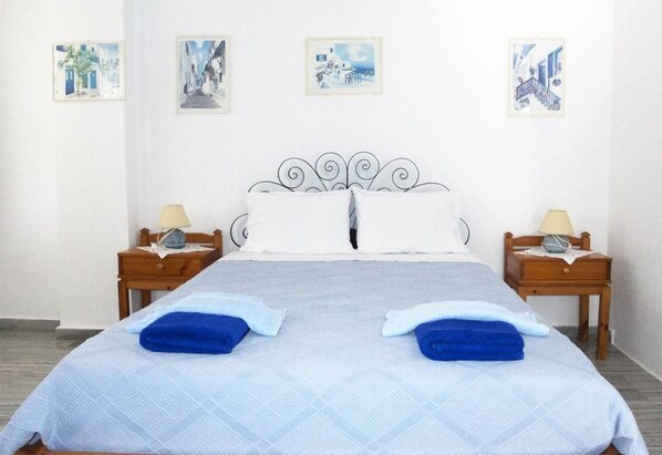 Een tweepersoonsbed met een wit-blauwe spreidek, twee kussens, twee blauwe handdoeken en een metalen hoofdbord. Er staan twee nachtkastjes bij, met lampen, en erboven hangen er fotolijstjes aan de muur.