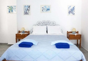 Cama de casal com colcha branca e azul, duas toalhas azuis e cabeceira de metal. Há duas mesas de cabeceira com abajures e uma parede com quatro quadros.