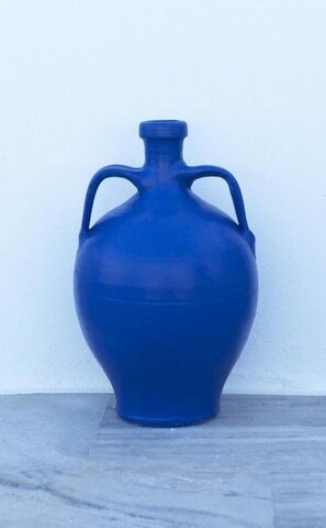 Um vaso cerâmico azul grande, com duas alças, colocado sobre um piso de azulejos, encostado em uma parede lisa.