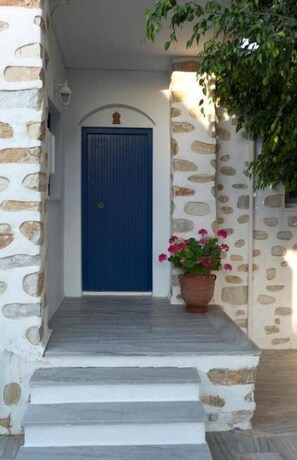 A stone-walled building with a blue door, a potted plant with pink flowers, and a small archway above the door. - Bungalows Marina (Paros)