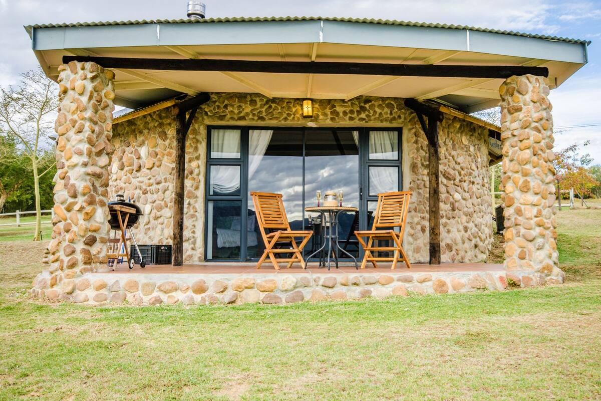 A stone building with a covered patio, two wooden chairs, a table, and a grill.