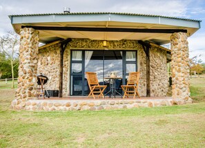 A stone building with a covered patio, two wooden chairs, a table, and a grill.