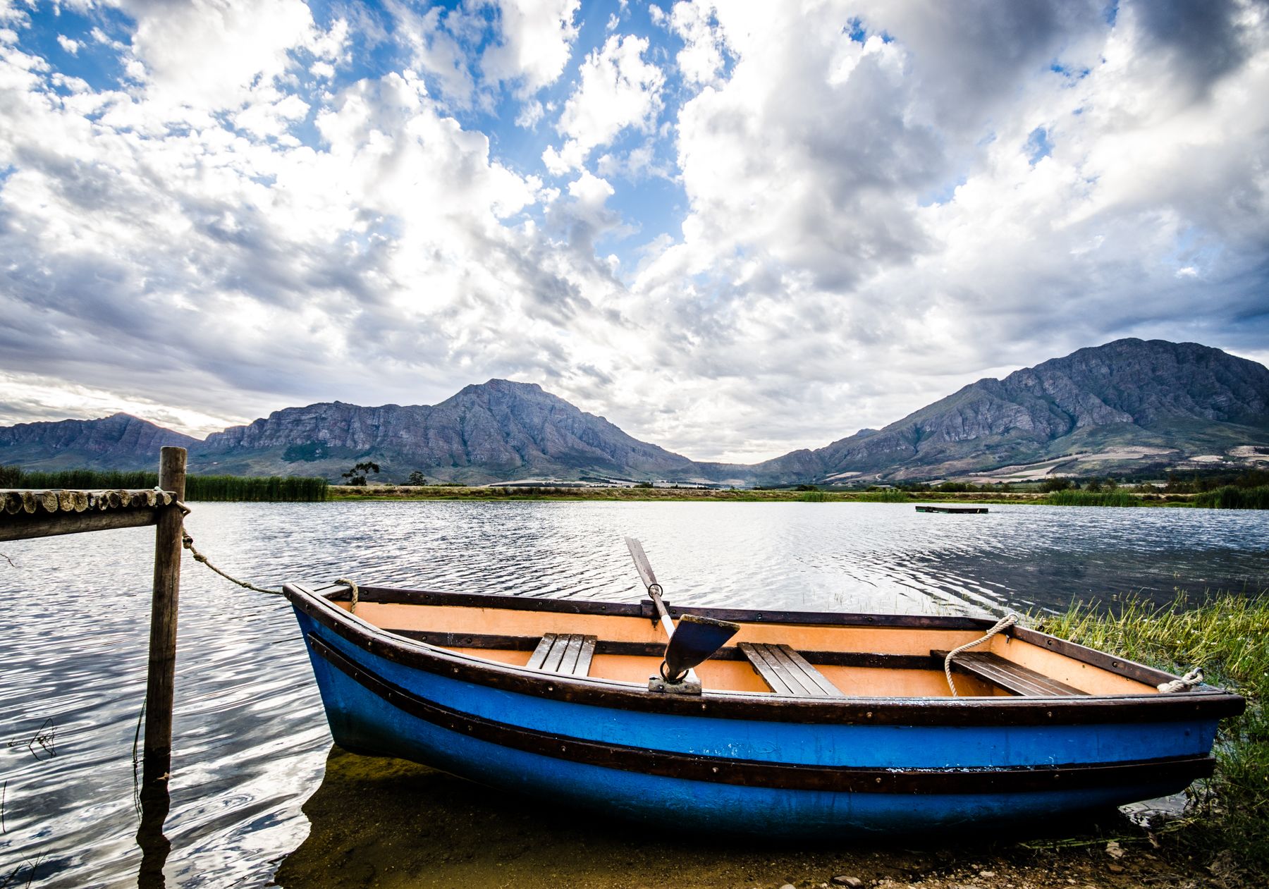 Un barco azul amarrado a un muelle, con un lago tranquilo y montañas al fondo.