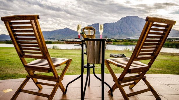 Two wooden chairs facing a table with a metal pitcher and glasses, set against a backdrop of a lake and mountains. - Reflections Guest Farm (Tulbagh)