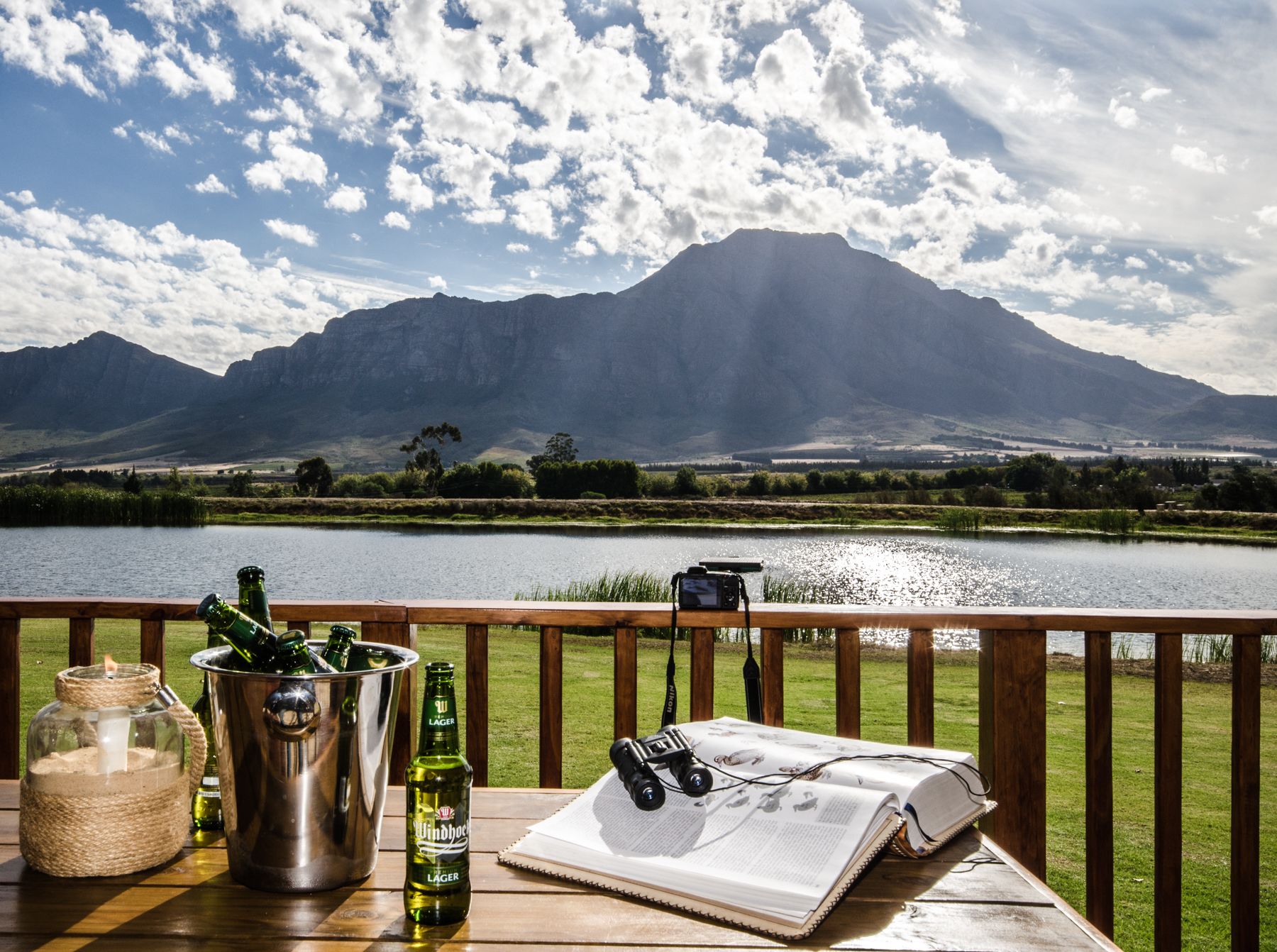 Una terraza de madera con una mesa donde hay una botella de cerveza, una cámara y un libro abierto. Se ve una montaña y un lago al fondo.