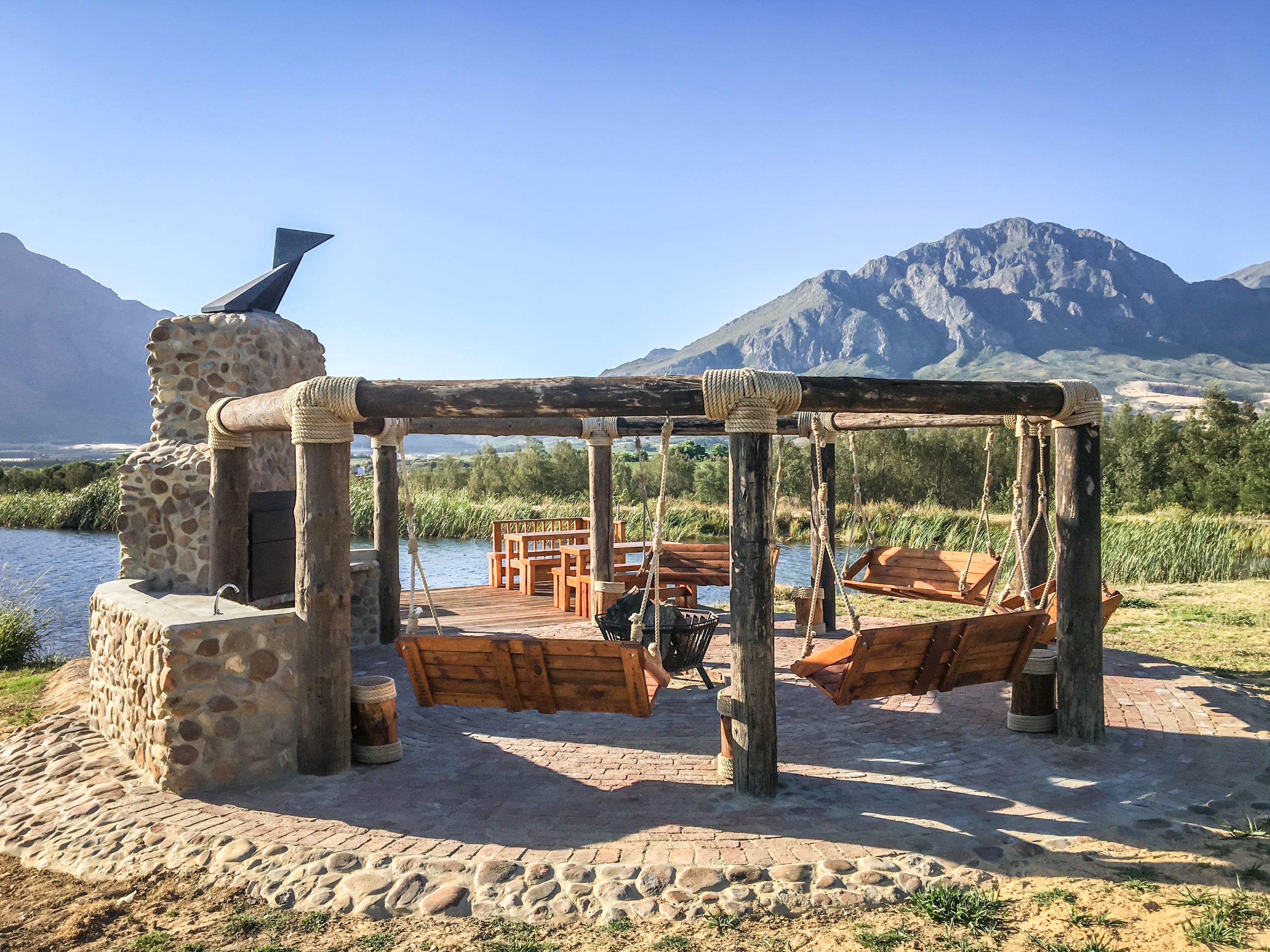 A rustic wooden swing set with benches, a stone wall, and a view of a river and mountains.