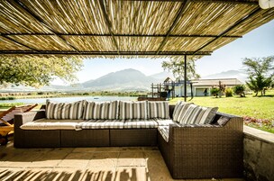 A covered outdoor seating area with a wicker sofa and striped cushions, overlooking a lake and mountains.
