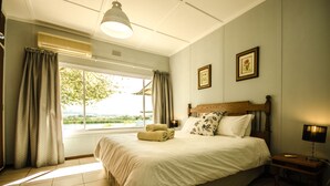 A bedroom with a large bed, a window with curtains, a bedside table with a lamp, and a wall-mounted air conditioner. - Reflections Guest Farm (Tulbagh)
