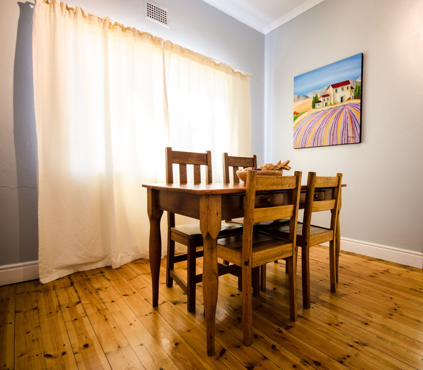 A dining area with a wooden table and chairs, a white wall with a painting, and a window with curtains.