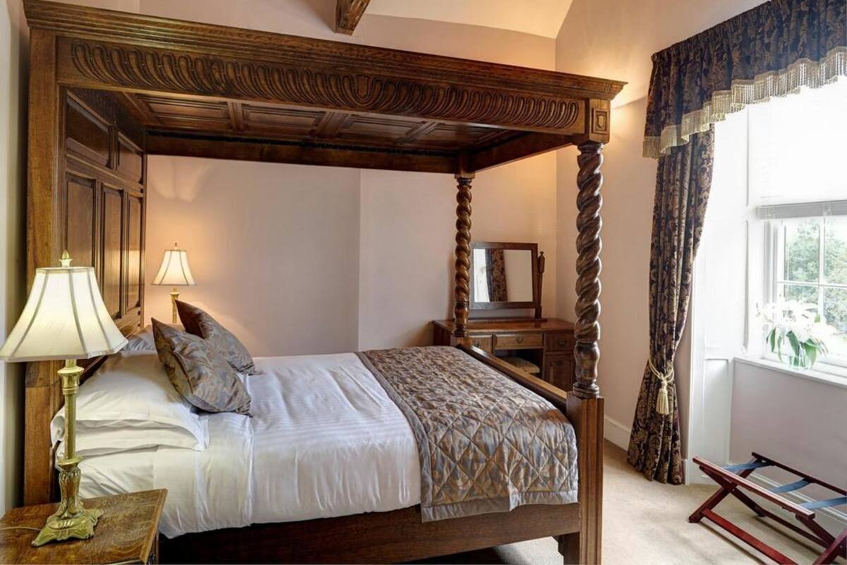 Un lit à baldaquin avec une tête de lit en bois, une table de chevet avec une lampe, un miroir et une fenêtre avec des rideaux.