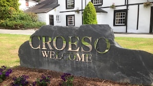 A welcome sign for Croeso with a stone base and metal letters, set against a backdrop of a white building with black trim and a well-maintained garden with purple flowers.