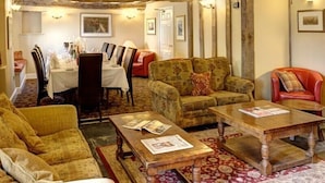 A rustic interior with exposed wooden beams, a dining area with a table set for four, and a seating area with two sofas and a red chair.