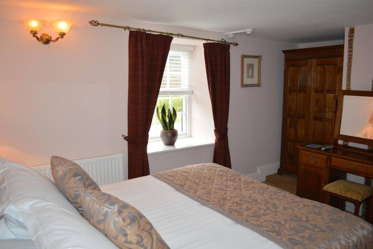 Une chambre à coucher comprenant un lit, une commode, une fenêtre avec des rideaux, une plante en pot et un luminaire fixé au mur.
