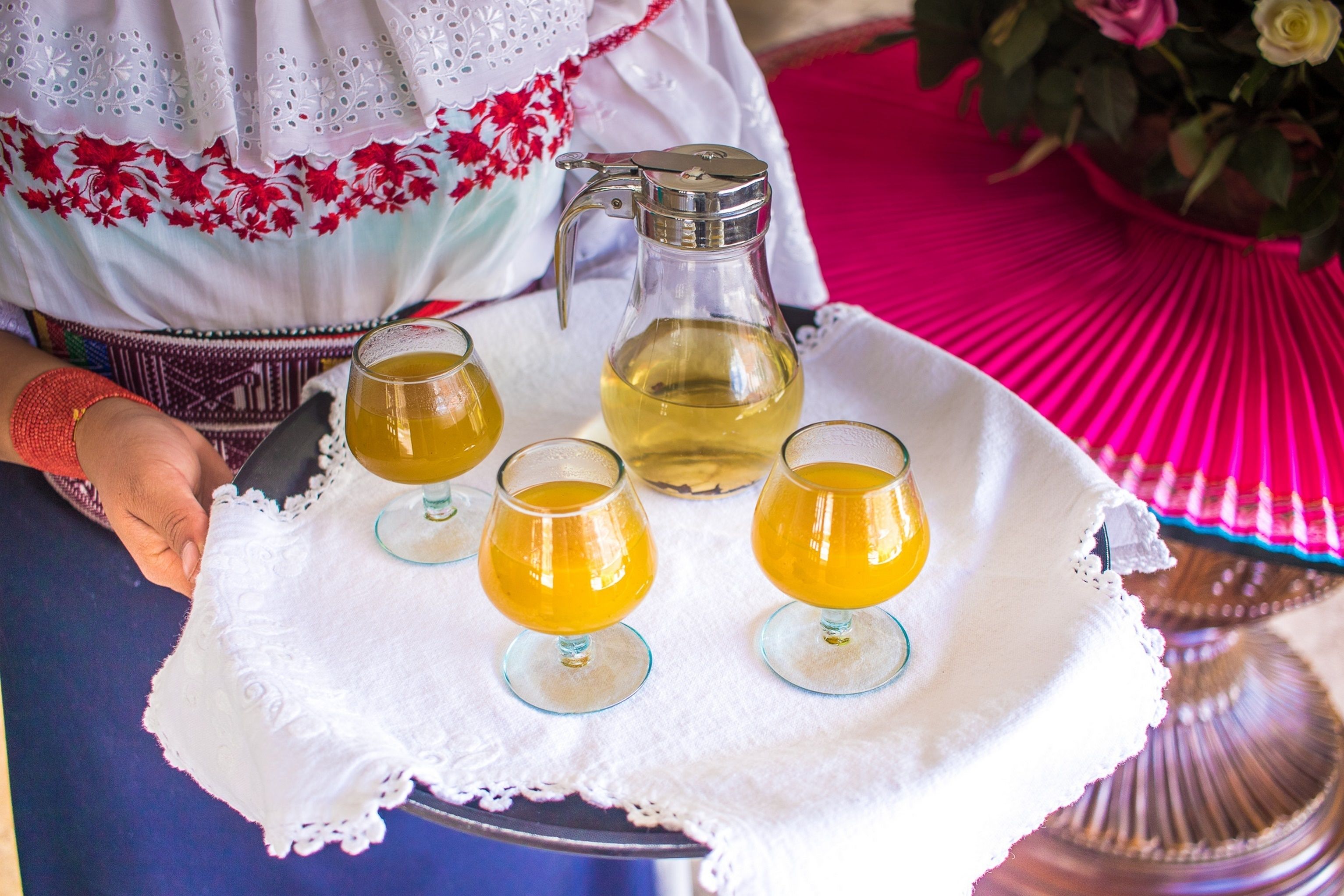 A tray with a pitcher and three glasses of a yellow beverage, placed on a white lace tablecloth.