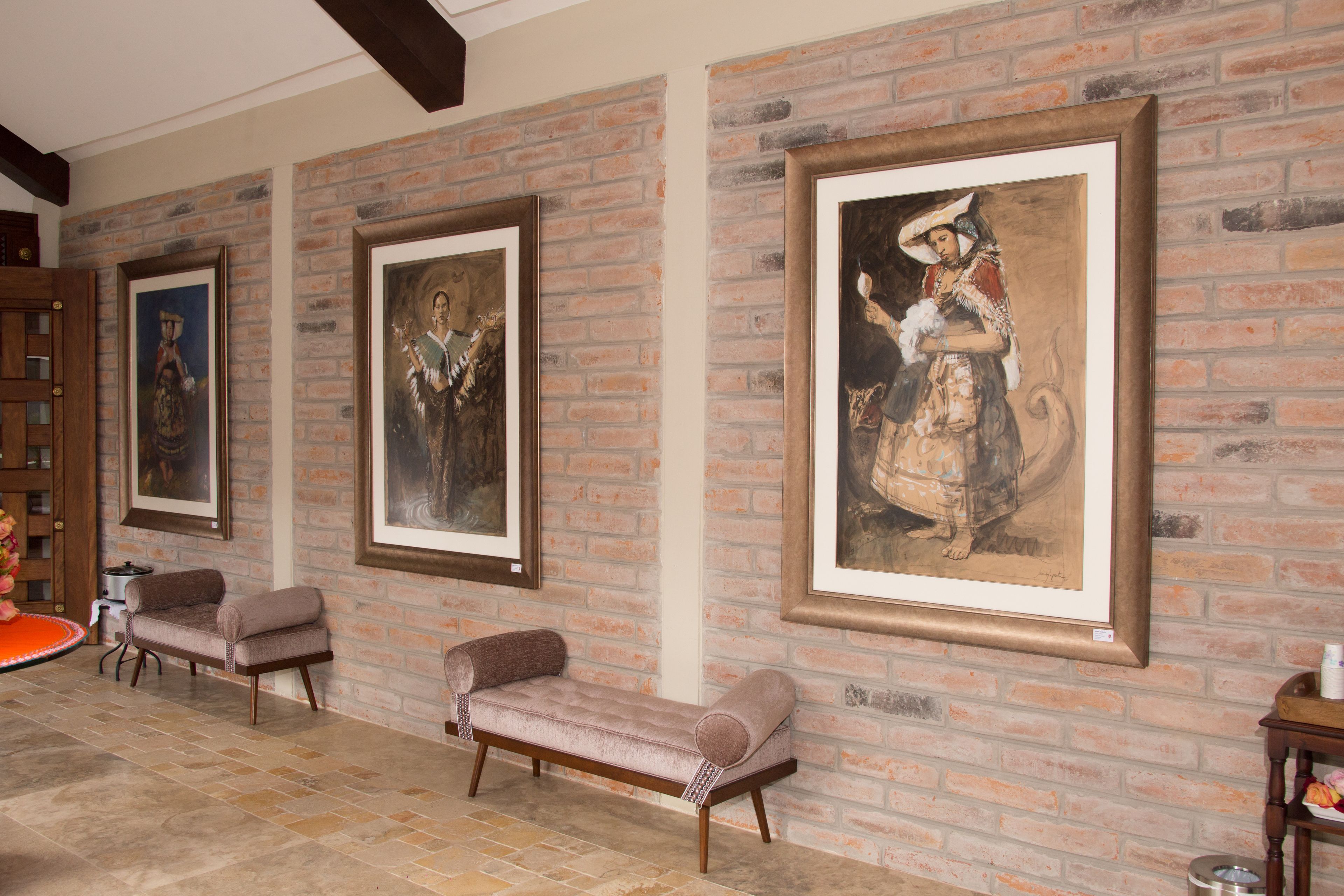 A room with a brick wall and three framed paintings. There are two upholstered chairs and a small table.