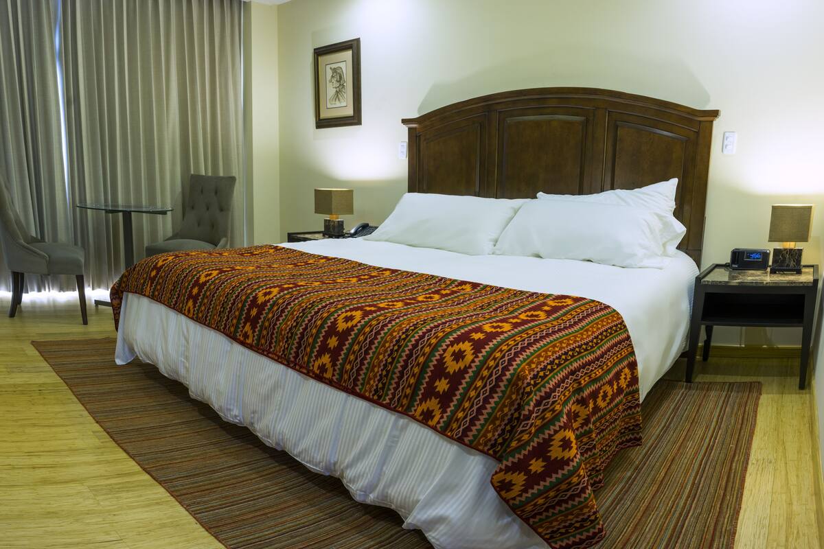 A hotel room with a large bed, a wooden headboard, a small table, a chair, and a nightstand with a lamp.