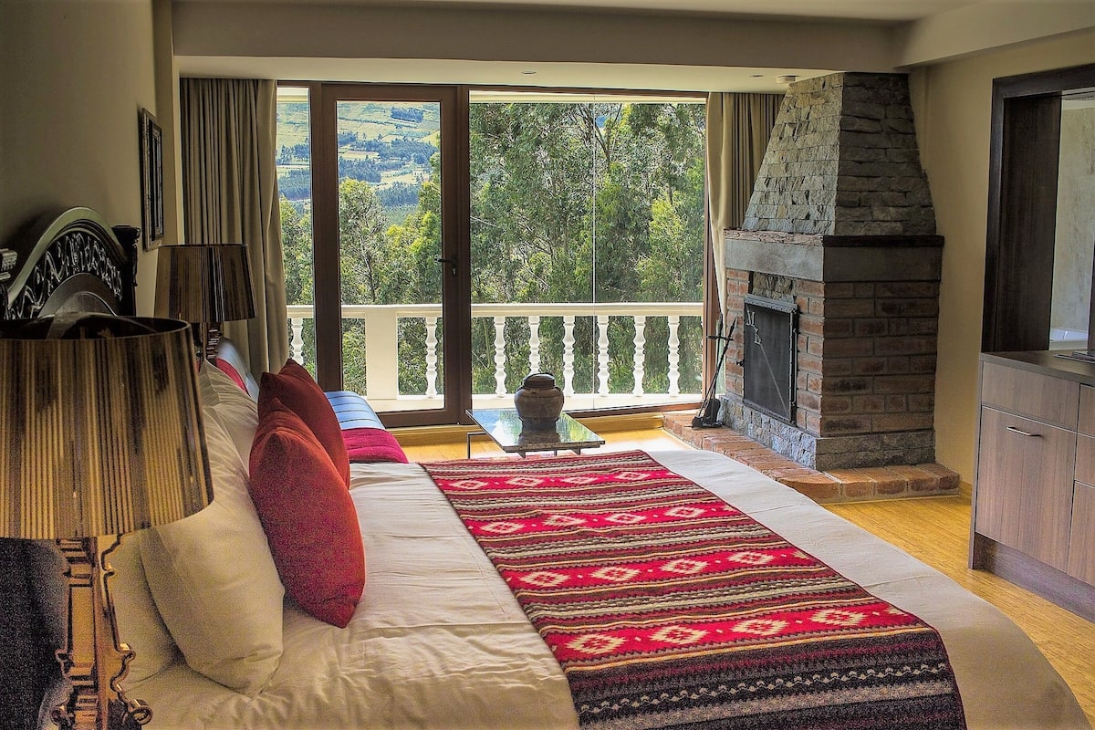 A bedroom with a large bed, a fireplace, a balcony with a view of the countryside, and a dining area with a table and chairs.
