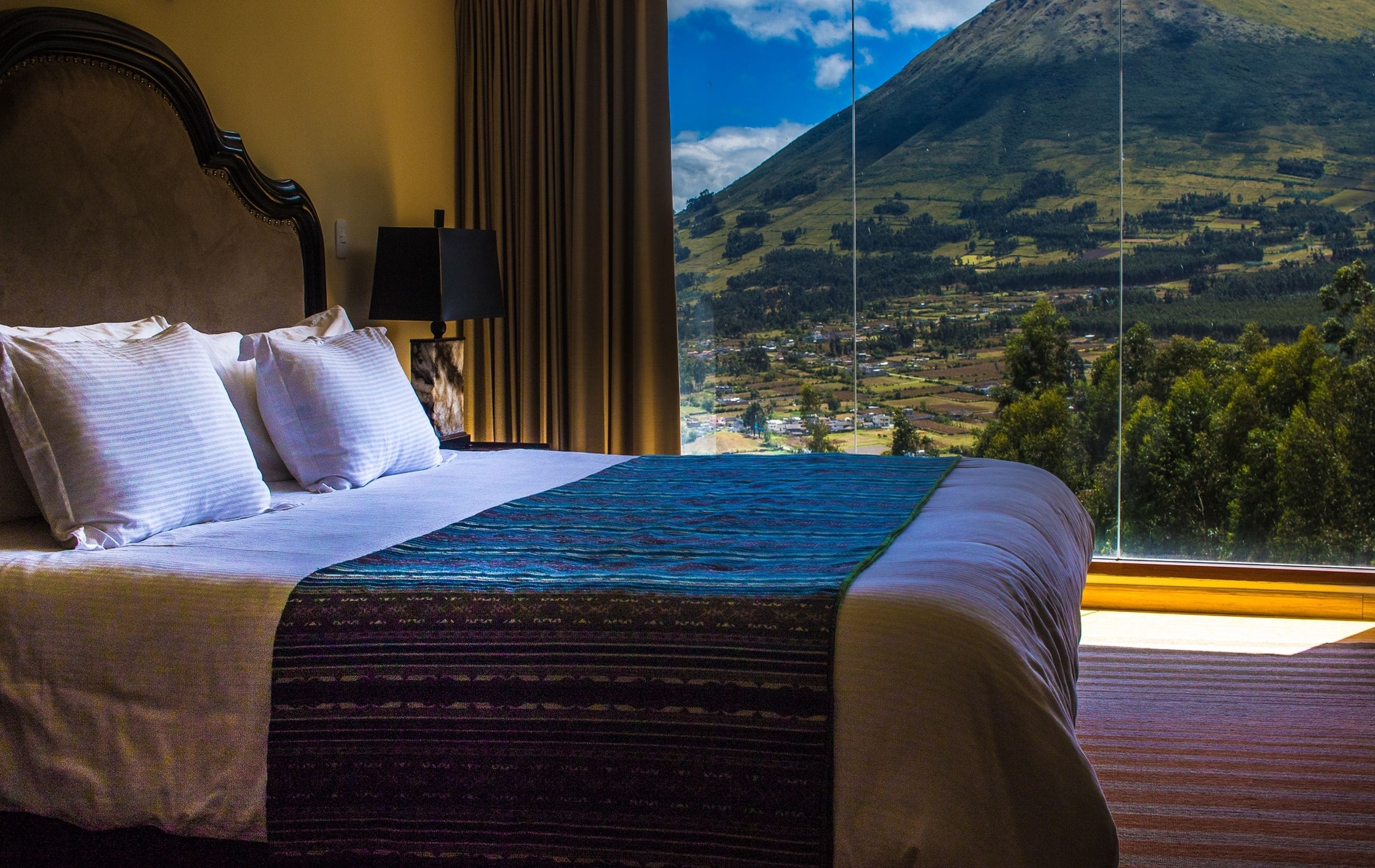 Una cama con sábanas blancas y una colcha estampada, una mesita de noche con una lámpara, y vistas de un paisaje montañoso a través de una ventana.