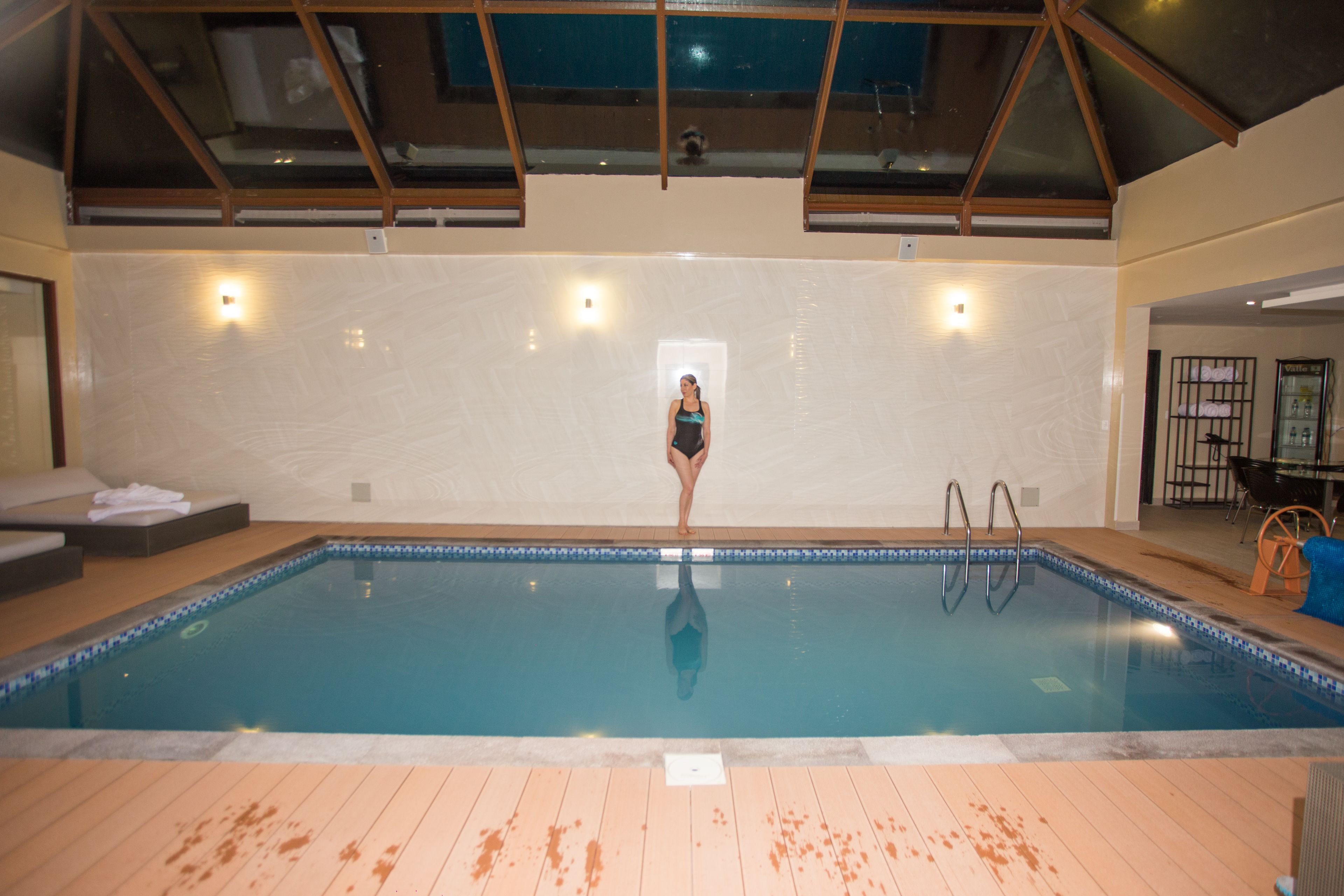 En inomhuspool med ett kaklat golv, ett glastak och en person som står vid poolkanten.