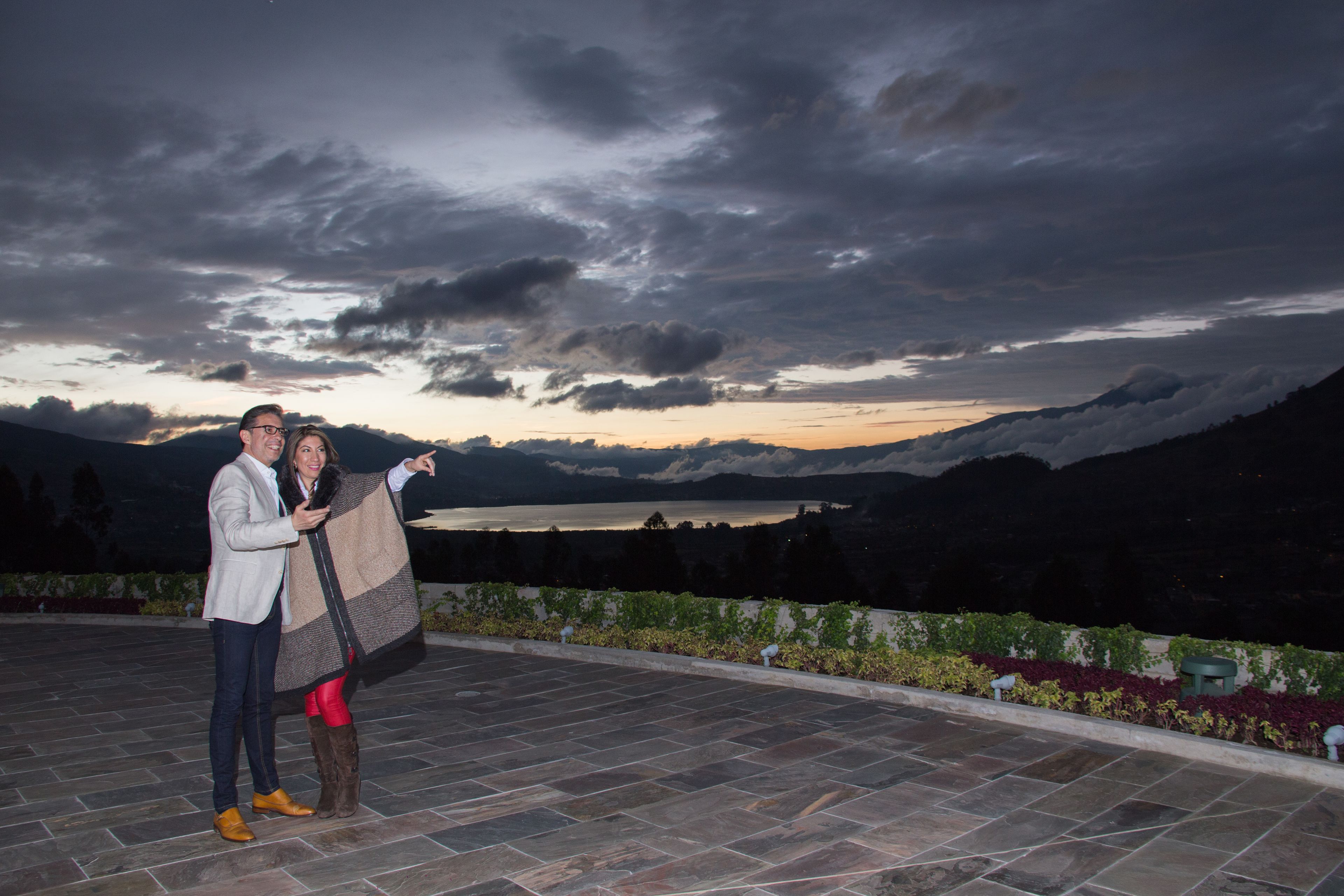 A couple on a terrace with a view of mountains and a lake at dusk.
