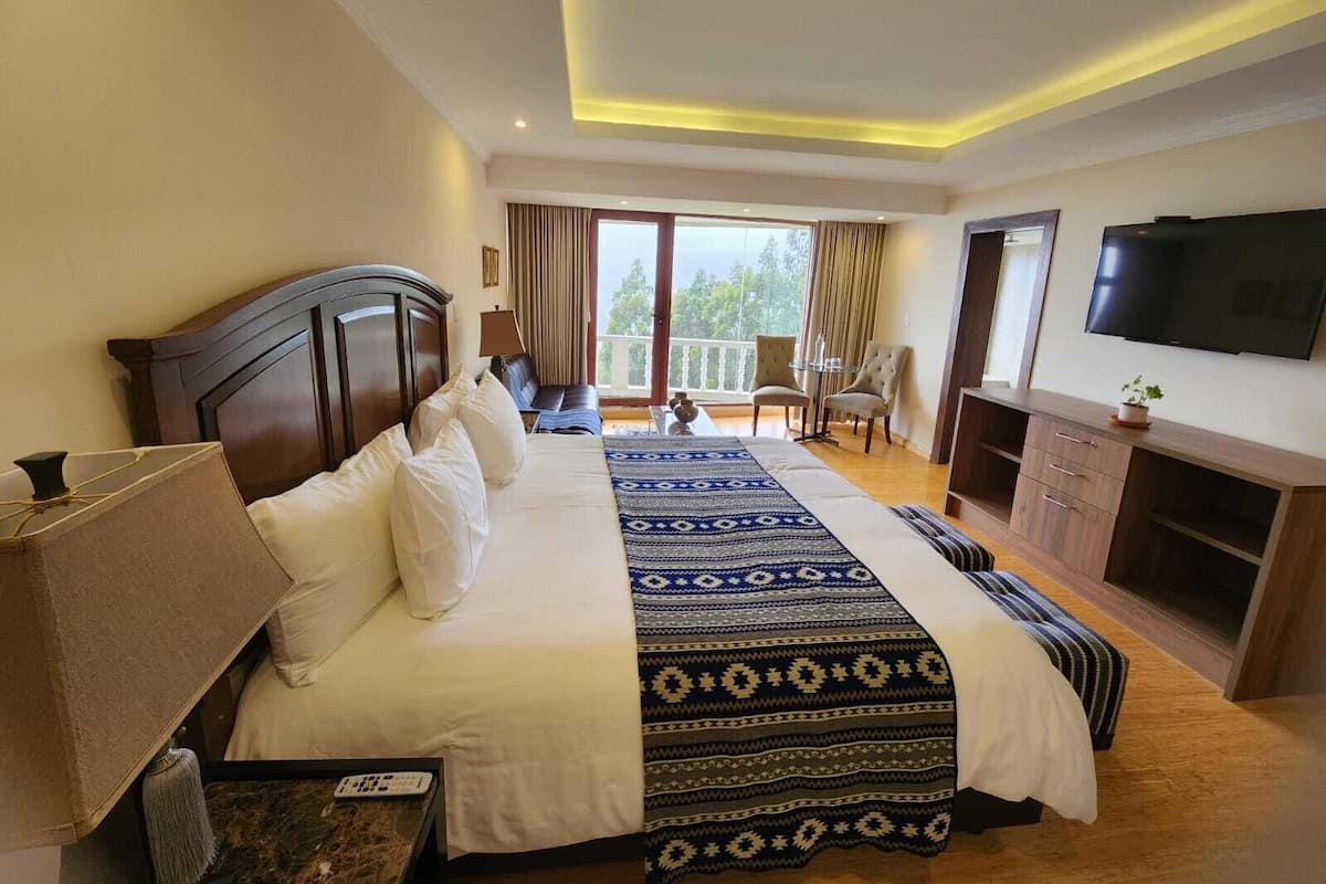 A hotel room with a large bed, a TV, a desk with a lamp, and a balcony with curtains.