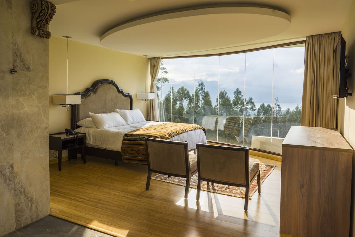 A spacious bedroom with a large bed, a desk, and a chair. There is a large window with a view of trees and mountains.