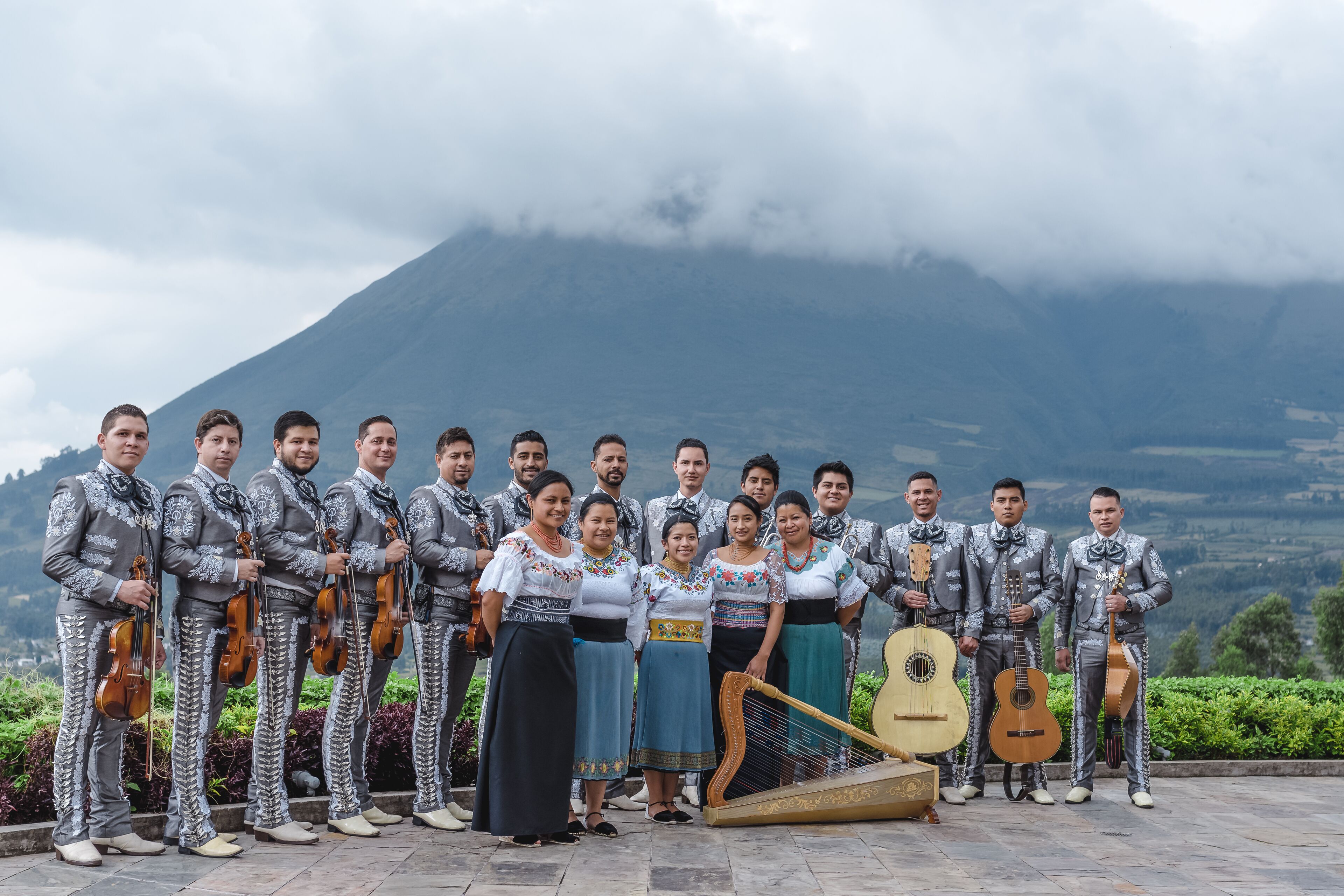 A group of musicians in traditional attire with musical instruments, standing in front of a mountain with clouds.
