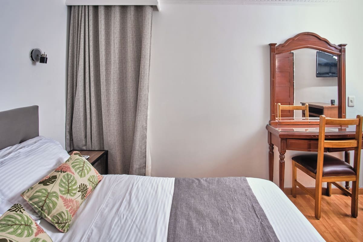 A hotel room with a bed, a desk, a chair, a mirror, and a window with curtains.