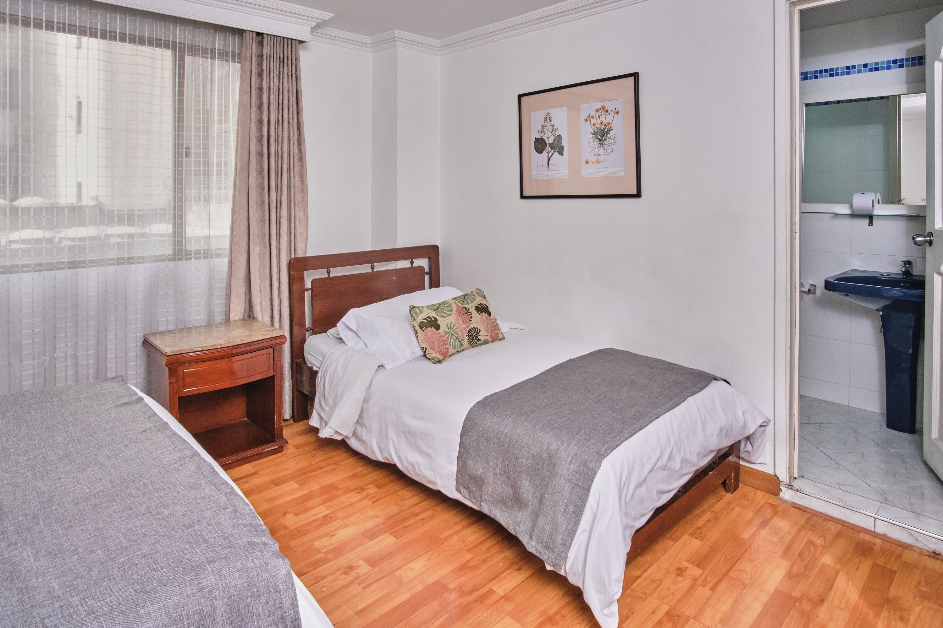 A bedroom with a wooden bed, a nightstand, a window with curtains, and a bathroom visible through an open door.