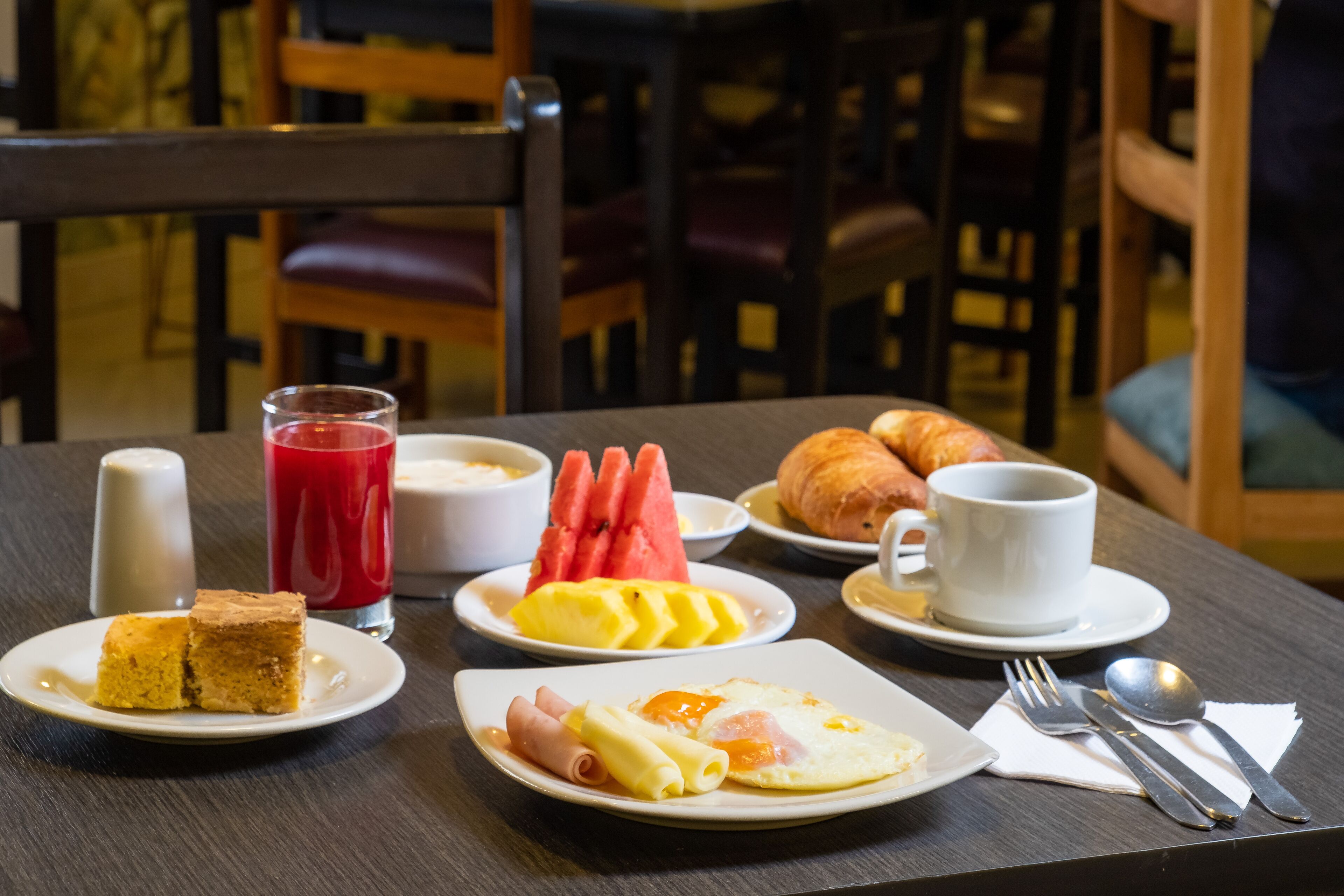 A breakfast table with plates of food, a glass of red juice, and breakfast items like eggs, ham, and croissants.