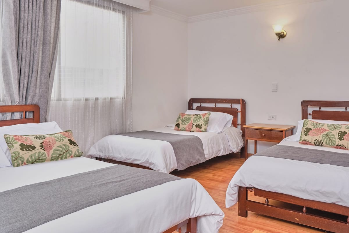 A hotel room with two single beds, each with a grey bedspread and a floral pillow. There is a wooden nightstand between the beds, and a window with sheer curtains.