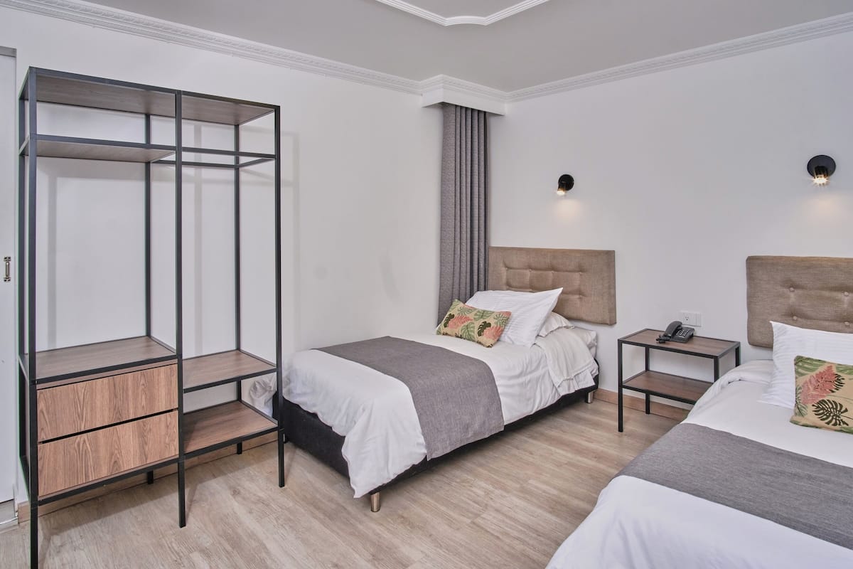 A hotel room with two beds, a wooden wardrobe, a nightstand, and a wall-mounted light fixture.