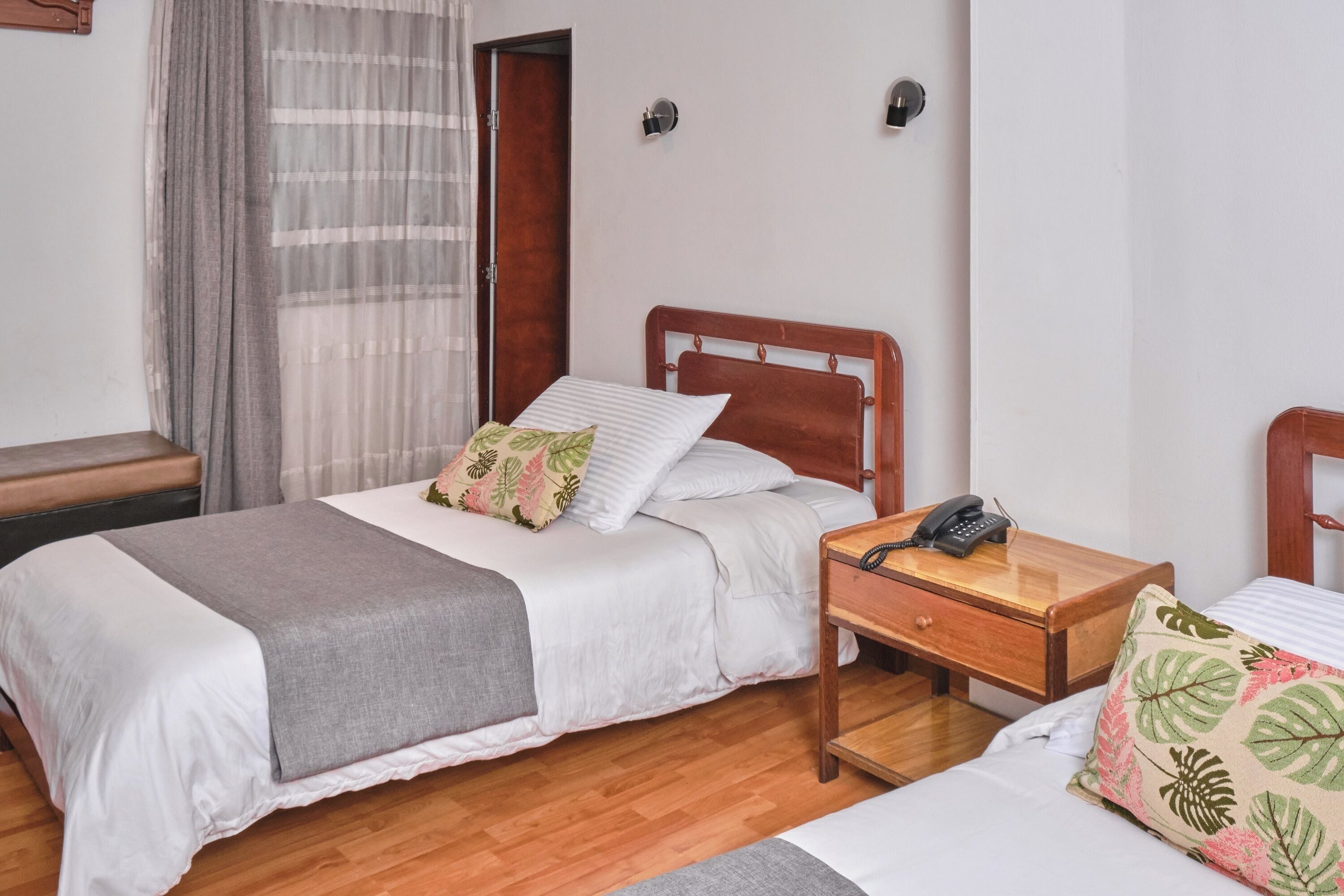 A double bed with a grey blanket, a wooden bedside table with a telephone, and a nightstand with a lamp.