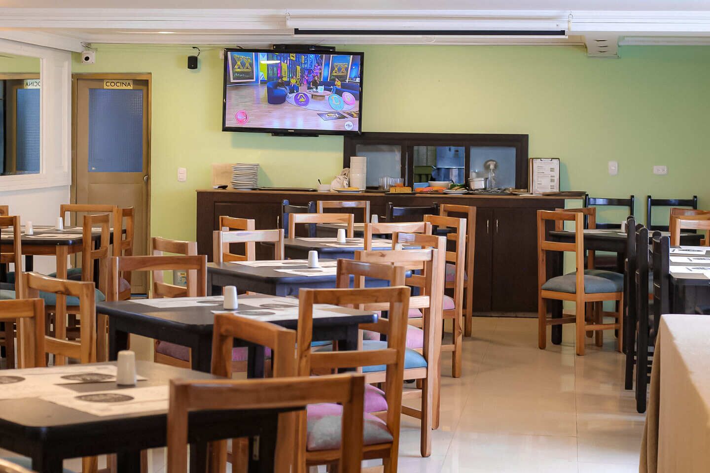 Un ristorante con diversi tavoli e sedie, una televisione montata a parete e una zona cucina con bancone ed elettrodomestici.