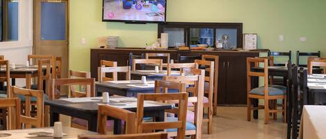 Un ristorante con diversi tavoli e sedie, una televisione montata a parete e una zona cucina con bancone ed elettrodomestici.